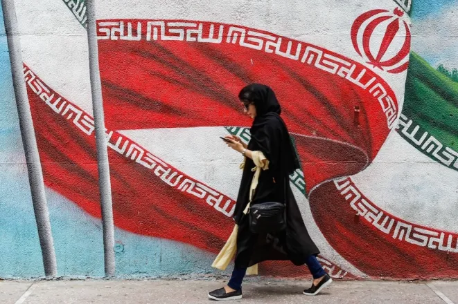 Perempuan muda Iran berjalan di depan mural bendera nasional, simbol dinamika gerakan politik Iran modern.
